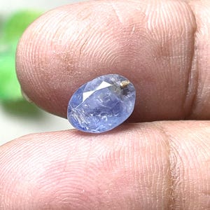 May include: An oval-shaped, faceted blue sapphire gemstone with visible internal inclusions and a translucent appearance. The stone displays varying shades of light to medium blue and features a polished surface with multiple geometric facets.