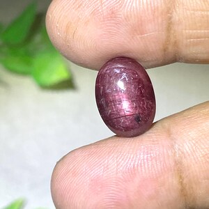 May include: An oval-shaped, deep red cabochon gemstone with a smooth, polished surface. The stone displays a translucent quality with visible internal inclusions and a subtle star-like reflection across its centre, showcasing a rich, dark crimson hue.
