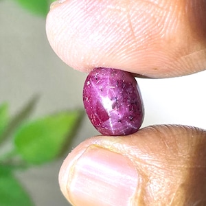 May include: A small, oval-shaped, deep magenta-colored gemstone with a smooth, polished surface and visible internal inclusions. The stone features a subtle star-like pattern reflecting light across its rounded, cabochon-cut top.