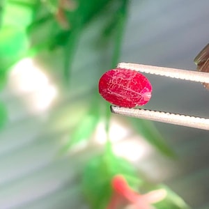 May include: A vibrant red, oval-shaped gemstone is held by silver tweezers against a blurred green background. The gem has a textured surface with visible internal patterns. The tweezers are held by a person.