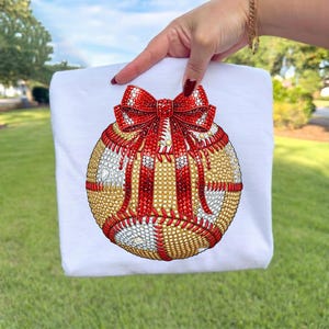 May include: White t-shirt featuring a baseball design with a red bow. The baseball is embellished with gold, red, and white rhinestones. The shirt is held up by a hand against a green grass background.