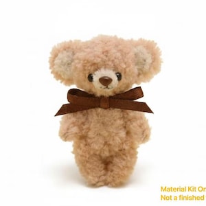 May include: A small, plush teddy bear with light brown fur and a dark brown ribbon tied around its neck. The bear has a small, dark brown nose and eyes. Text at the bottom reads "Material Kit Only Not a finished toy."