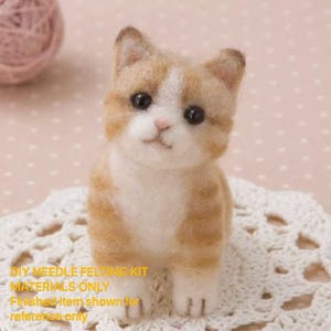 May include: A handmade, needle-felted kitten figurine in shades of tan and white, with black eyes and a pink nose. The kitten is sitting on a white, lacy doily. The image includes text that reads "DIY NEEDLE FELTING KIT MATERIALS ONLY Finished item shown for reference only."