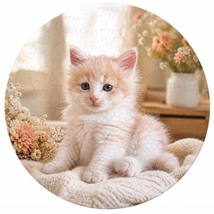 以下が含まれることがあります： 青い目のふわふわとした薄いオレンジと白の子猫が描かれた丸いジグソーパズル。子猫はクリーム色の毛布の上に座っており、花瓶や横にドライフラワーが飾られています。パズルは柔らかく温かみのある画像です。