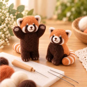 May include: Two needle-felted red panda figures made of wool. One figure stands with arms raised, while the other sits. Both have orange and dark brown fur with striped tails. A wooden felting tool and loose wool roving are on a white foam pad.