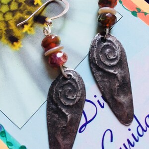 May include: Silver earrings with a spiral design and brown and pink gemstone beads. The earrings are on a white background with the text "Divina Guilda".