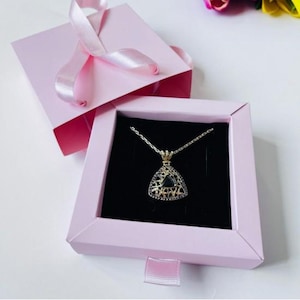 May include: A gold-colored pendant necklace with a triangular black stone, presented in a pink gift box with a satin ribbon. The necklace is displayed on a black velvet background.