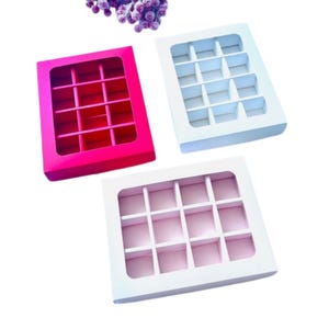 Puede incluir: Tres cajas de caramelos con compartimentos divididos. Una es rosa brillante, otra es blanca y la tercera también es blanca con el interior rosa. Cada caja tiene una ventana transparente en la parte superior.