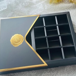 May include: A gray jewelry box with a gold trim and a gold emblem on the lid. The box is divided into multiple compartments. The box is open, revealing the interior compartments.