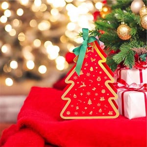 Puede incluir: Una caja de regalo roja en forma de árbol de Navidad con detalles dorados y una cinta verde. La caja está decorada con símbolos festivos dorados. La caja está sobre una superficie roja cerca de un árbol de Navidad y regalos envueltos.