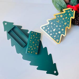 Puede incluir: Un juego de cajas de regalo con temática navideña. El juego incluye una caja verde oscuro en forma de árbol con tapa a juego y una caja rectangular más pequeña. Ambas cajas están decoradas con iconos dorados navideños.