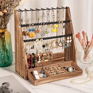May include: A wooden jewelry organizer stand with a top horizontal bar for hanging necklaces and earrings, featuring two black metal racks with holes. The base includes a rectangular tray with three compartments lined with beige fabric for rings.