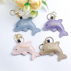Puede incluir: Cuatro llaveros con forma de delfín en colores pastel: azul, morado, rosa y beige. Cada llavero tiene un anillo dorado y una correa negra. Los nombres "Leo", "Miro", "Maren" y "Taren" están impresos en cada delfín.