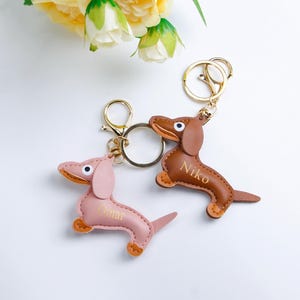 Peut inclure: Deux porte-clés en cuir en forme de teckel. L'un est rose avec le nom "Omar" et l'autre est marron avec le nom "Niko". Les deux porte-clés ont des anneaux et des fermoirs en métal doré. Des fleurs jaunes sont en arrière-plan.