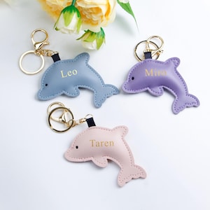 Puede incluir: Tres llaveros con forma de delfín en colores pastel: azul, morado y rosa. Cada llavero tiene un cierre dorado y una correa negra. Los nombres "Leo", "Mina" y "Taren" están impresos en cada delfín.