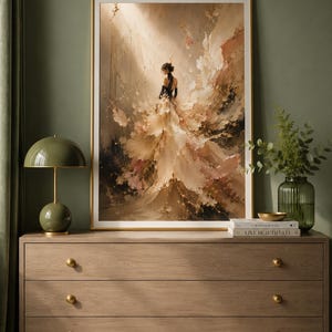 May include: An abstract art print in a gold frame depicts a woman in a flowing gown. The artwork features a palette of beige, brown, and pink hues, with black accents. The print is displayed above a wooden dresser with gold knobs, complemented by a green lamp and vase.