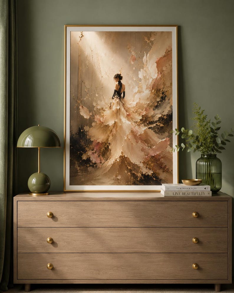 May include: An abstract art print in a gold frame depicts a woman in a flowing gown. The artwork features a palette of beige, brown, and pink hues, with black accents. The print is displayed above a wooden dresser with gold knobs, complemented by a green lamp and vase.