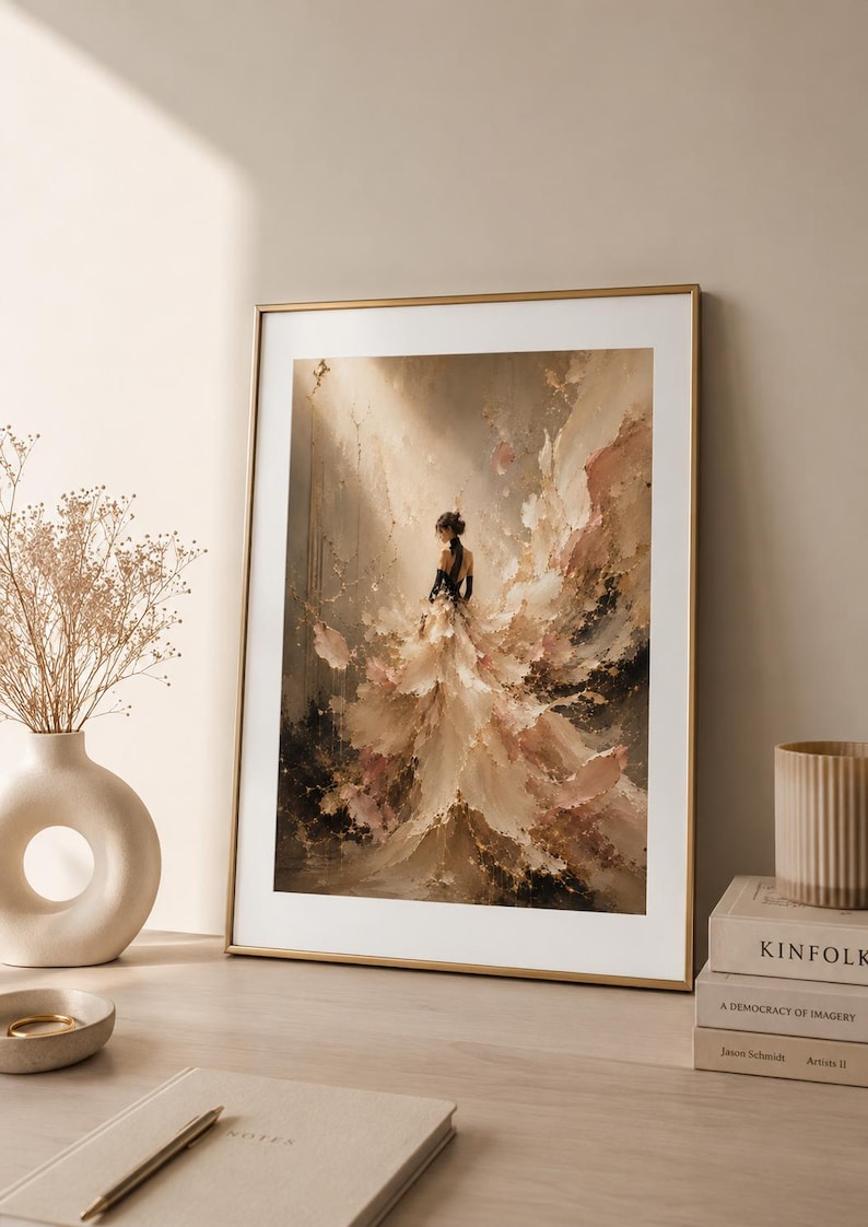 May include: An abstract painting in a gold frame depicts a figure in a flowing gown. The artwork uses a palette of beige, brown, and black, with a focus on texture and light. The framed art is displayed on a wooden surface.