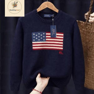 May include: Navy blue sweater featuring an American flag design on the front. The flag has a blue square with white stars and red and white stripes. The letters "RL" are below the flag. The sweater has a crew neck and a tag that says "POLO".