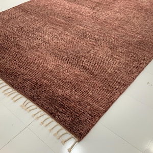 May include: A rectangular, textured rug in a warm, reddish-brown hue. The rug features a woven design with a fringe of cream-colored tassels along one edge. The rug is placed on a white tiled floor.