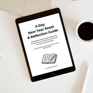 Pode incluir: Um tablet preto exibe o título "3-Day New Year Reset & Reflection Guide" com texto descrevendo uma jornada de escrita. Um laptop, xícara de café, caneta e objeto decorativo também são visíveis.