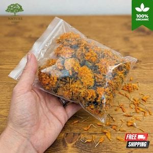 Puede incluir: Una bolsa de plástico transparente resellable llena de flores de caléndula secas de color naranja. La bolsa contiene numerosos cabezales de flores enteros y fragmentados con pétalos de colores naranja y amarillo vibrantes.