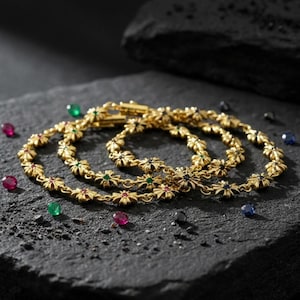 May include: Three gold bracelets with a floral design, each adorned with red, green, and blue gemstones. The bracelets are displayed on a dark, textured surface, with scattered gemstones around them.