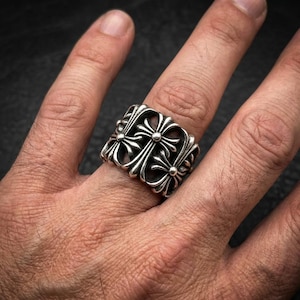 以下が含まれることがあります： 繰り返し十字模様のシルバーリング。リングは厚みがあり、詳細で装飾的なパターンがあります。十字はバンドの周りに均等に配置され、大胆で人目を引くアクセサリーを形成しています。リングの幅は約2cmです。