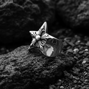 May include: A silver-colored ring with a star-shaped design. The star features a central circular element surrounded by ornate floral patterns. The ring is set against a dark, textured background, highlighting its metallic sheen.