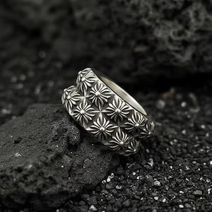 以下が含まれることがあります： 繰り返し花柄模様のシルバーリング。リングは光沢のある金属製で、詳細なデザインが施されています。リングは、暗く質感のある表面に表示されています。