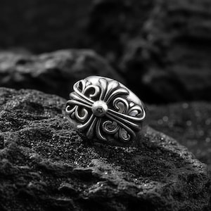 May include: A silver ring with an ornate, symmetrical design featuring a central sphere and stylized floral elements. The ring is set against a dark, textured background, highlighting its intricate details and metallic sheen.
