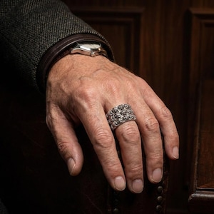 May include: A silver ring with a detailed, geometric design is displayed on a hand. The ring has a textured surface and a wide band. The person is wearing a watch with a silver band and a dark jacket.