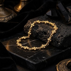 May include: A gold-colored bracelet featuring a repeating floral cross design. The bracelet is displayed on a black marble surface, with a dark background. The jewelry is made of metal and has a clasp closure.