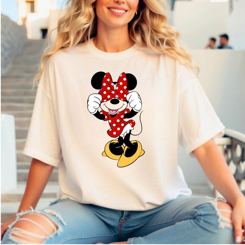 May include: White t-shirt featuring a cartoon Minnie Mouse design. Minnie is depicted in a red dress with white polka dots, a matching bow, and yellow shoes. The design is centered on the shirt.