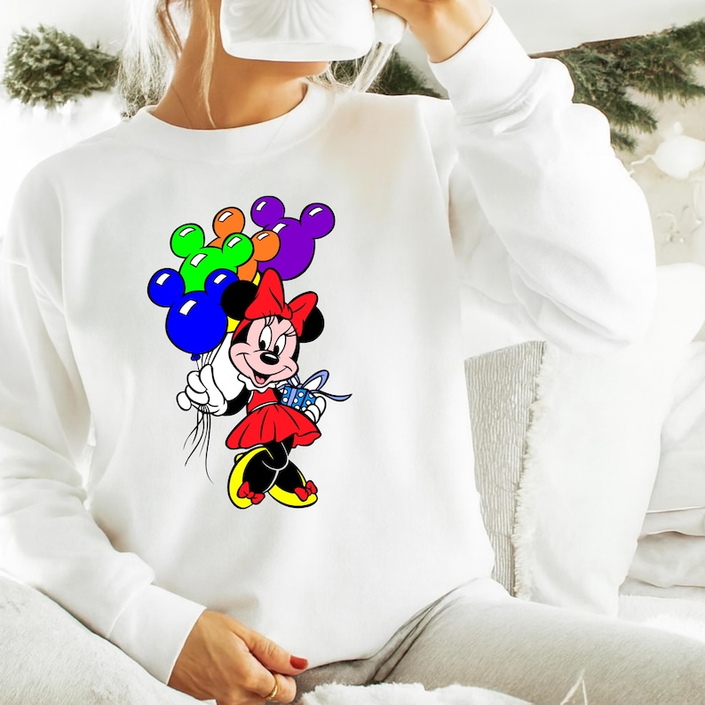 May include: White sweatshirt featuring a colorful Minnie Mouse graphic. Minnie Mouse is holding a bouquet of Mickey Mouse-shaped balloons in blue, green, orange, and purple. She is wearing a red dress, a red bow, and yellow shoes.