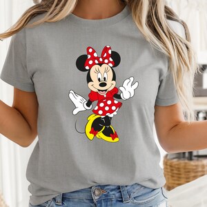 May include: A gray t-shirt featuring a cartoon illustration of Minnie Mouse. Minnie is depicted in a red dress with white polka dots, a matching bow, and yellow shoes. The design is centered on the shirt.