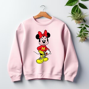 May include: A light pink sweatshirt featuring a cartoon Minnie Mouse design. Minnie Mouse is wearing a red top with a yellow skirt and shoes. The design includes a red bow and the letter "M".