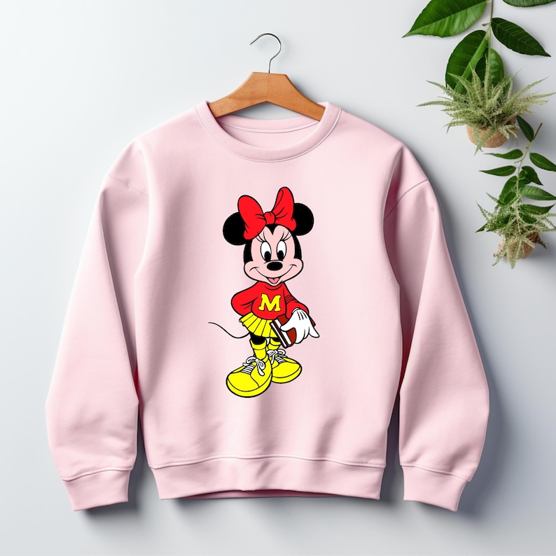 May include: A light pink sweatshirt featuring a cartoon Minnie Mouse design. Minnie Mouse is wearing a red top with a yellow skirt and shoes. The design includes a red bow and the letter "M".
