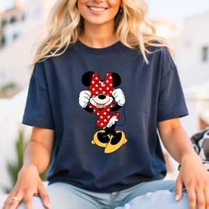 May include: Navy blue t-shirt featuring a Minnie Mouse graphic. The cartoon character is depicted in a red dress with white polka dots, a matching bow, and yellow shoes. The design is centered on the shirt.