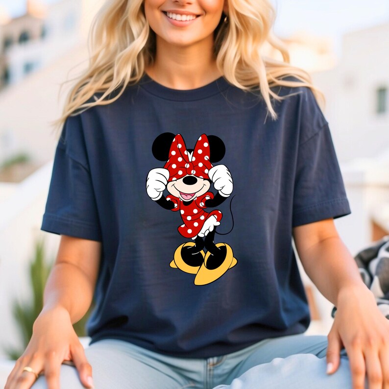 May include: Navy blue t-shirt featuring a Minnie Mouse graphic. The cartoon character is depicted in a red dress with white polka dots, a matching bow, and yellow shoes. The design is centered on the shirt.
