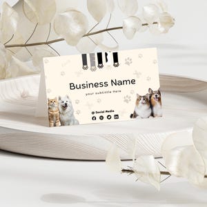 May include: A cream-colored folded business card featuring a pattern of paw prints and bones. The card displays the text Business Name, your subtitle Here, and @ Social Media with social icons, alongside images of two cats and two dogs.
