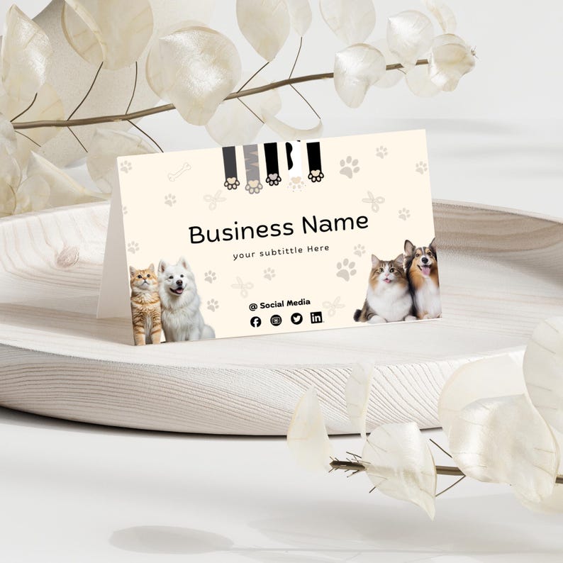 May include: A cream-colored folded business card featuring a pattern of paw prints and bones. The card displays the text Business Name, your subtitle Here, and @ Social Media with social icons, alongside images of two cats and two dogs.