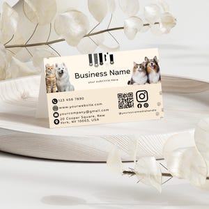 May include: A rectangular business card with a cream background featuring images of two cats and two dogs. The text reads Business Name, your subtitle Here, contact details, a QR code, and social media icons with the handle @yoursocialmediahandle.