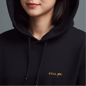 Peut inclure: Sweat à capuche noir avec l'inscription "STILL ME" en lettres dorées. Le sweat à capuche est doté d'une capuche à cordon de serrage et d'une coupe ample. L'image montre le haut du corps d'une personne portant le sweat.