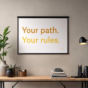 Könnte beinhalten: Gerahmter Druck mit dem Text "Your path. Your rules." in goldfarbenen Buchstaben. Der Druck hängt über einem Holzschreibtisch mit einer Lampe, Büchern und dekorativen Gegenständen. Eine grüne Pflanze in einer Vase befindet sich links.