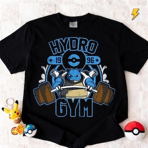 Puede incluir: Camiseta negra con un gráfico azul y marrón de un personaje Blastoise levantando pesas, con las palabras "HYDRO GYM 1996". Un llavero de Pikachu y una Pokeball son visibles.