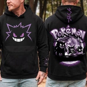 Puede incluir: Sudadera con capucha negra con un gráfico de personaje de dibujos animados púrpura en la parte delantera y trasera. El diseño frontal muestra la cara de un personaje, mientras que la parte posterior muestra la palabra "Pokemon" y otros personajes.