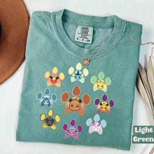 Op de afbeelding: Lichtgroen T-shirt met een pootafdrukontwerp met verschillende stripfiguren. Het shirt heeft een ronde hals en korte mouwen. Het merklabel "Comfort Colors" is zichtbaar op de bovenrug van het shirt. Een zilveren ketting met een hanger is over het shirt gedrapeerd.