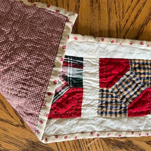 May include: A patchwork quilt runner with a red, white, and blue color scheme. The runner is made of different fabrics, including plaid, gingham, and polka dot. The runner is trimmed with a white fabric with a floral print.
