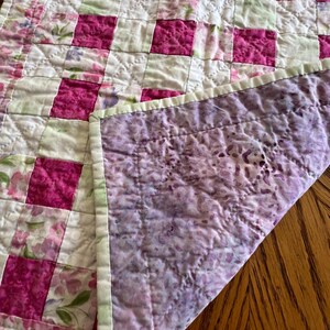 May include: A handmade quilt with a patchwork design. The quilt features alternating squares of vibrant fuchsia and floral patterned fabric. The reverse side is a mottled purple fabric. The edges are finished with a light green binding.